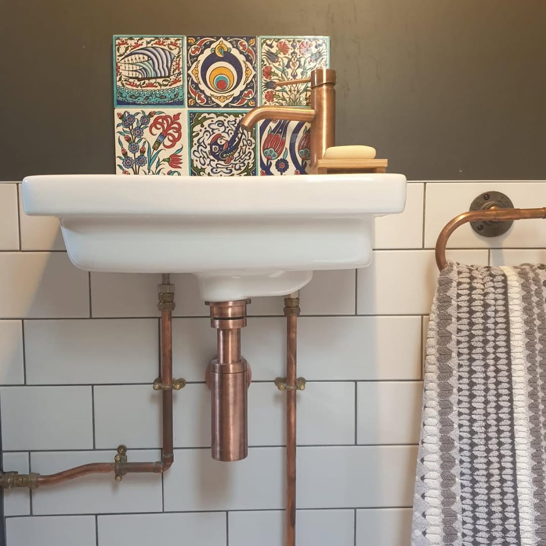 Close-up of copper tap and exposed copper pipes with hand-painted decorative tiles behind basin