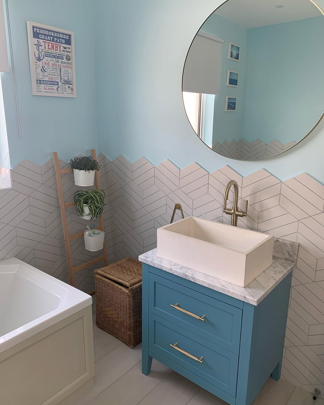 Bathroom vanity area with blue cabinet, marble countertop, large round mirror, and herringbone tile splashback